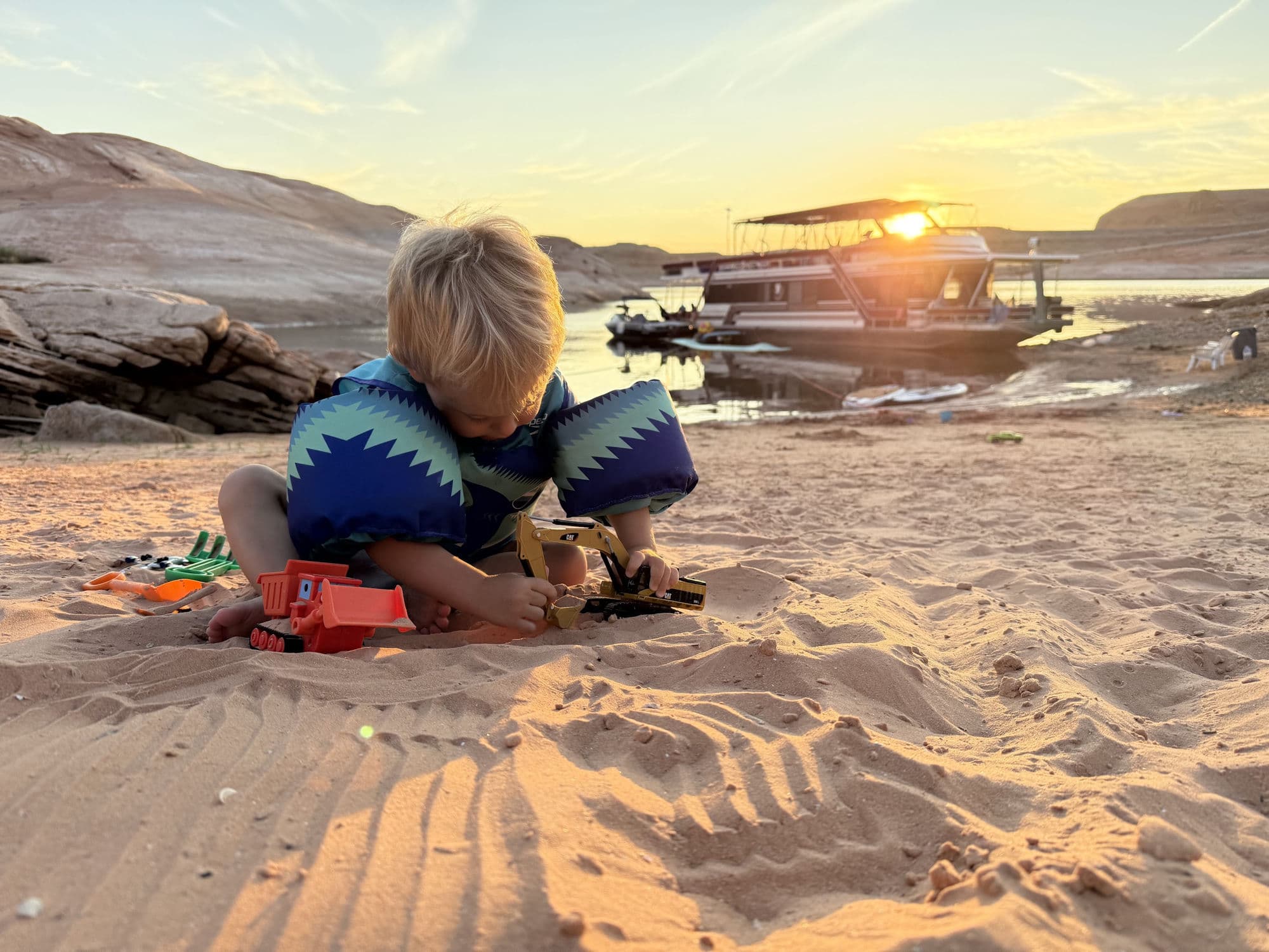 A child plays in the sand at Lake Powell at sunset, with a houseboat in the background